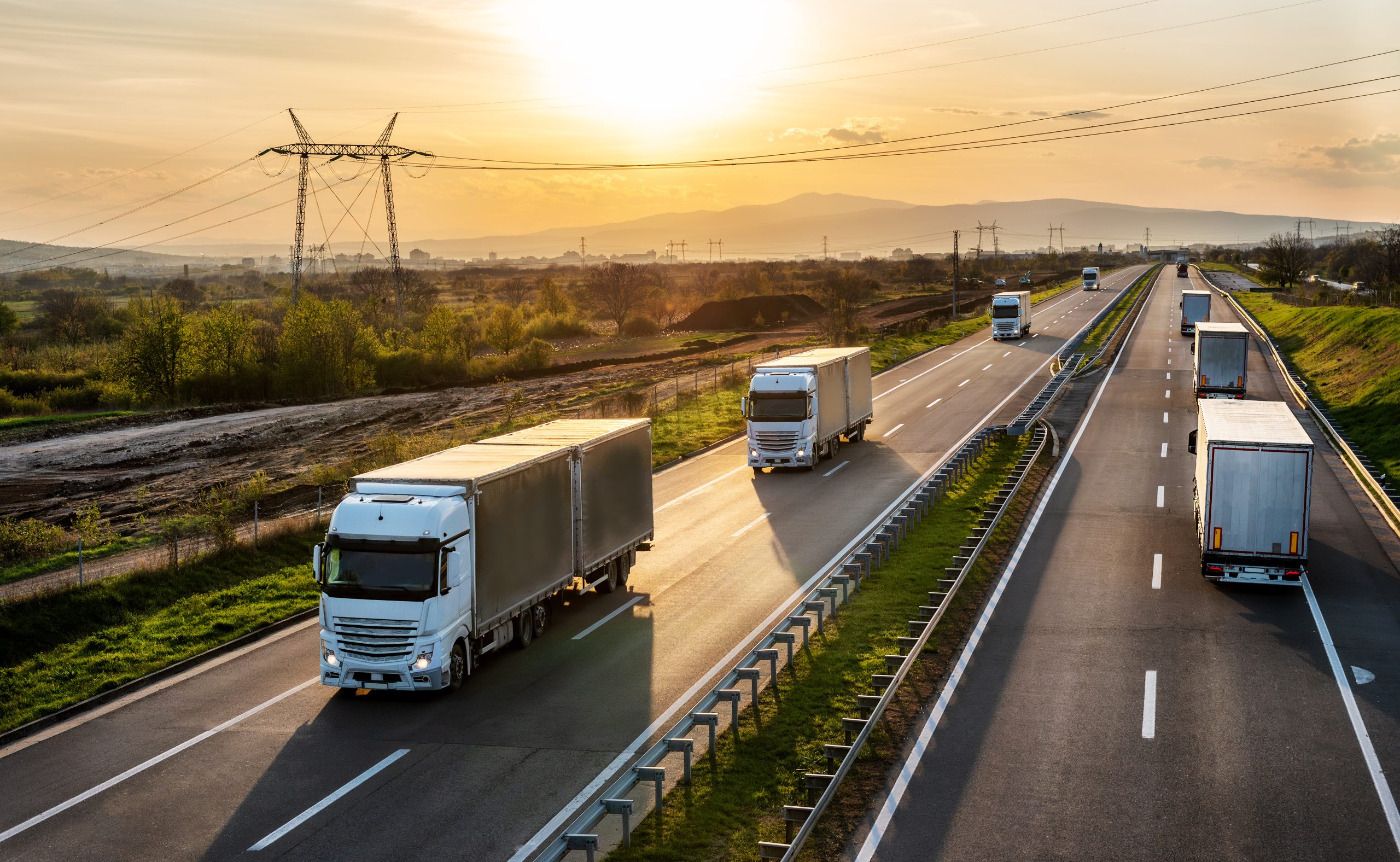 Big white transportation trucks with grey containers passing other trucks on a highway at sunset. Cargo transportation concept - freight service, on a highway in both ways