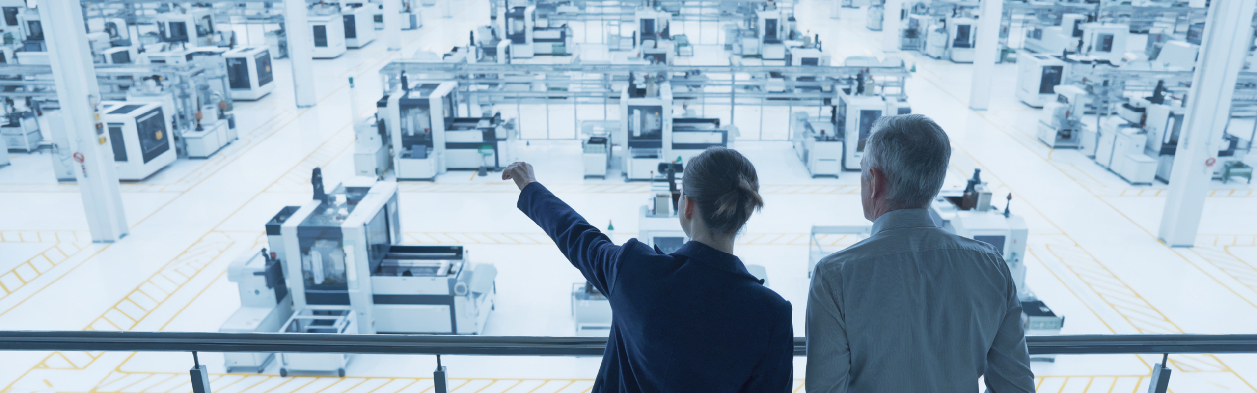 Two people in business attire overlook a vast automated factory floor filled with rows of manufacturing machines.